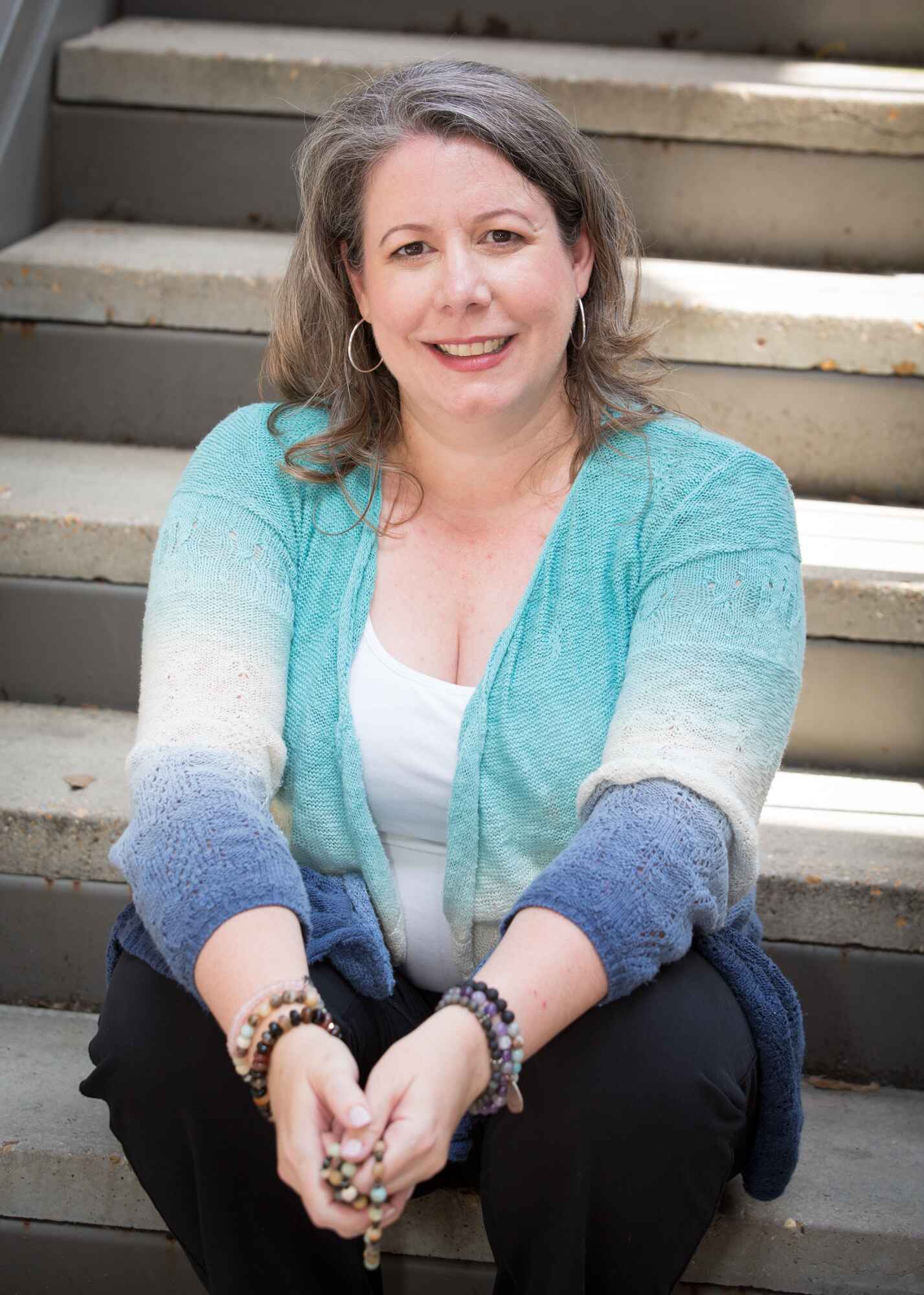 Woman in blue cardigan sitting on stairs with mala beads in hands.